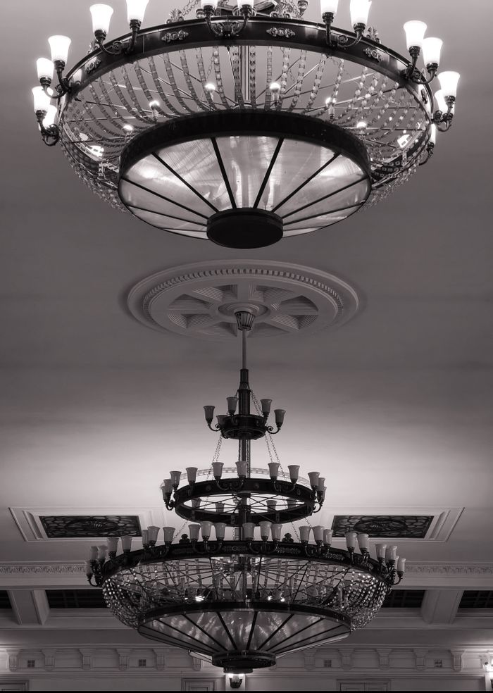 Black and white photo of two ornate vintage chandeliers in an elegant hallway with intricate ceiling design.